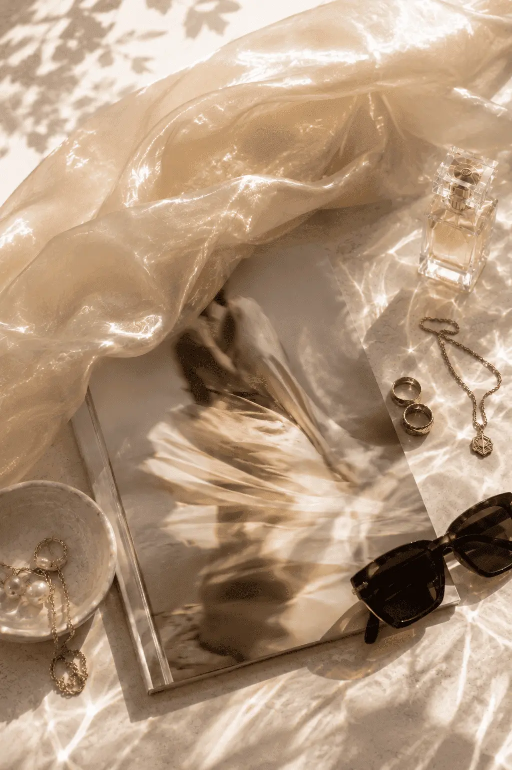Sheer champagne silk, a perfume bottle, gold jewellery and sunglasses arranged on marble in dappled sunlight